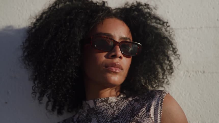 A woman with voluminous curly hair wears sunglasses and leans against a white wall