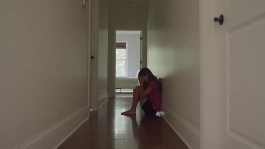 A teenage girl is with a back on a wall then sits on a shiny wood floor in a long white hallway her head rests on her knees an open doorway is visible at the end of the hall
