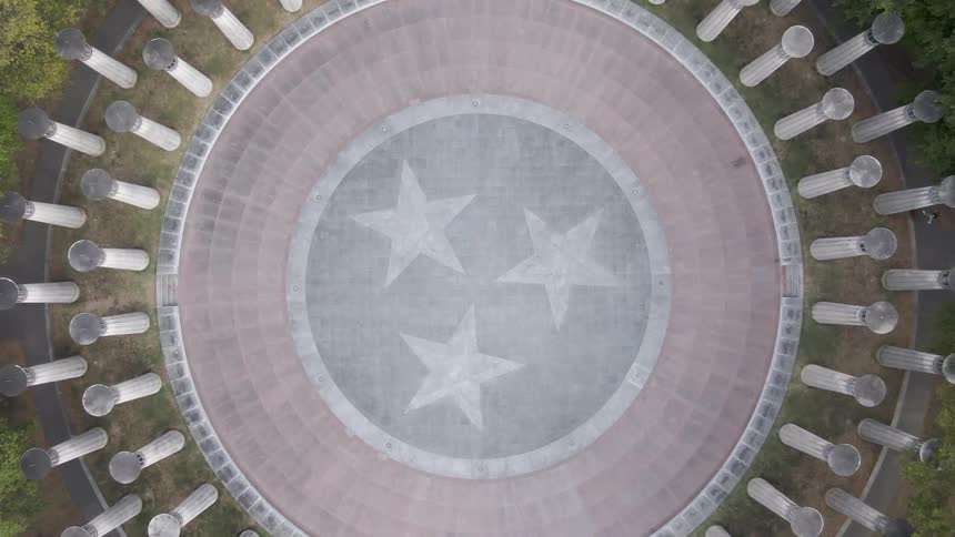 Aerial view of a paved circular clearing in a park surrounded by pillars with spherical lights on top