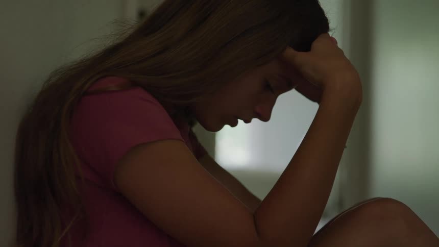 A close dark side view shows a sad long haired girl wearing a pink shirt sitting alone and resting her head on her hand in a dimly lit space