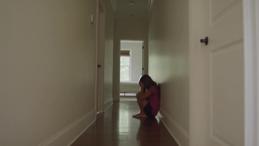 A girl with long hair wearing a pink shirt and black shorts slides down a light hallway wall to sit alone on the hardwood floor with her knees tucked to her chest