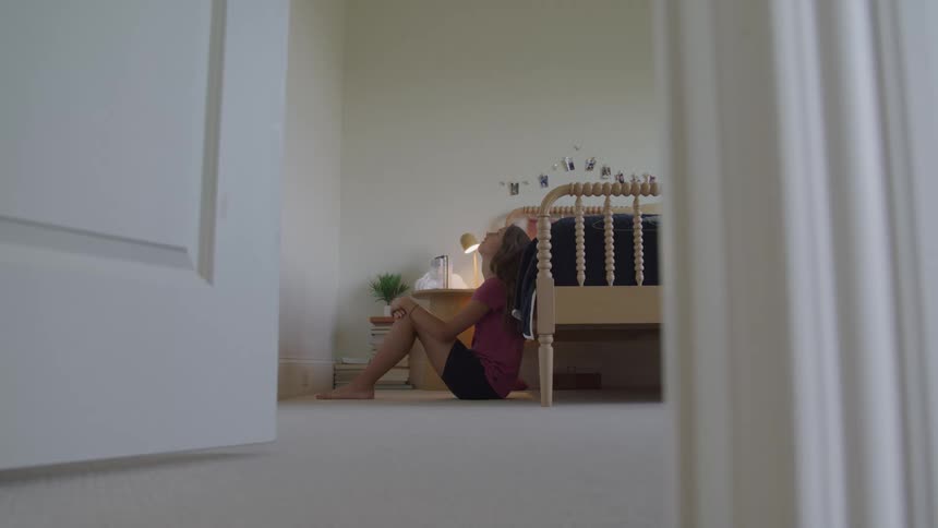 The video shot through an open doorway shows a young woman sitting alone on the floor of a bedroom with her back against the bedshe is sitting in a distressed pose with her head resting on her knees suggesting sadness stress or isolation