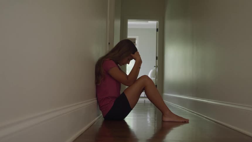 A young long haired girl wearing a pink shirt and black shorts sits alone crying with her head in her hands on the shiny hardwood floor of a long dimly lit hallway