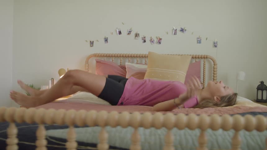 A girl with long brown hair wearing a pink shirt and black shorts walks into a bright bedroom and falls backward onto a bed with a colorful blanket