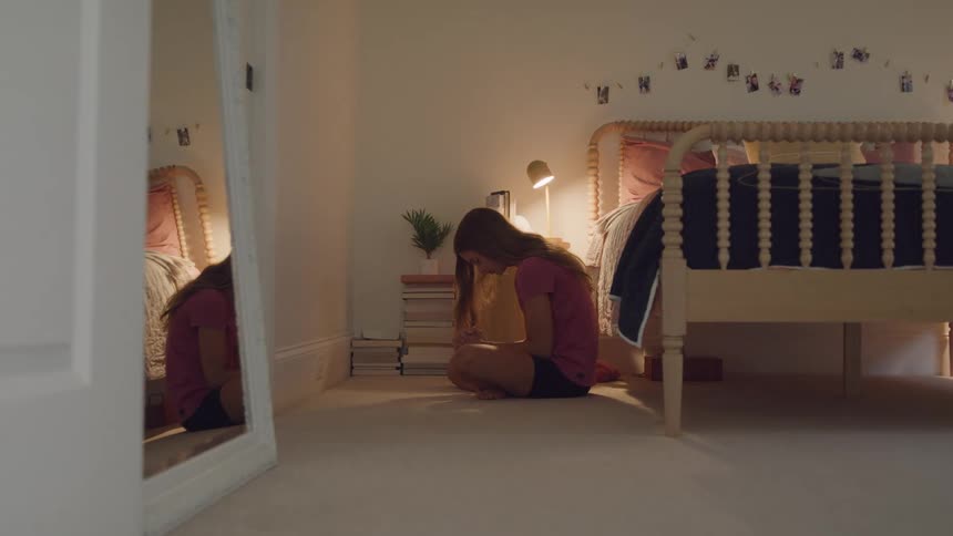 A sad teenage girl is sitting cross legged by the bed in her bedroom