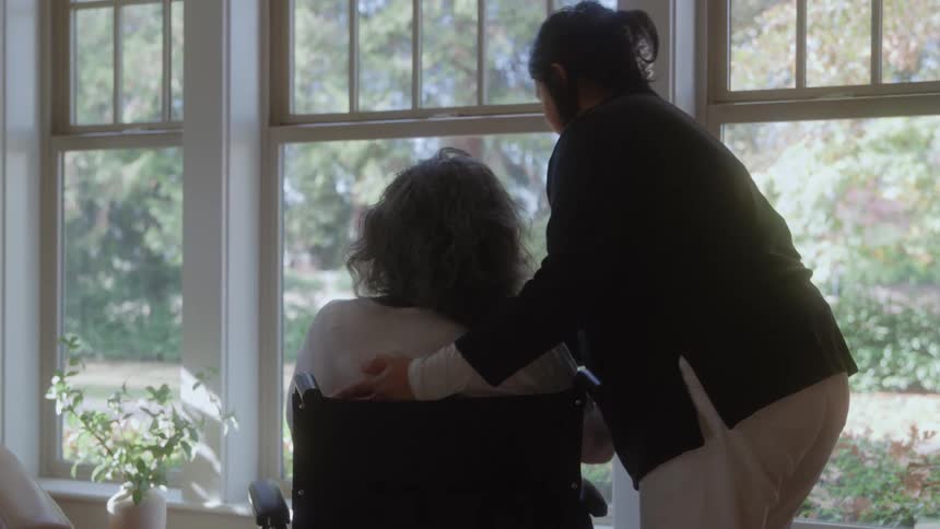 The video is a mediumclose shot of an adult in a dark shirt hugging an older person in a wheelchair from behindthe embrace is tender and takes place indoors near a large window suggesting a caring moment between family or caregiver and patien