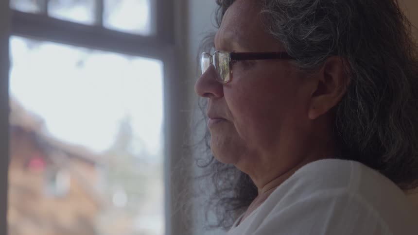 An older woman with glasses and wavy graying hair looks thoughtfully out a window her face illuminated by the natural light
