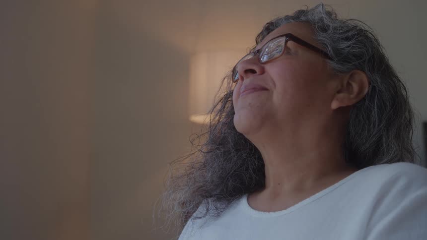 The video is a closeup profile shot of an older woman with long gray wavy hair and glassesshe is looking toward a bright source of light coming from the right giving her face a contemplative or peaceful expression
