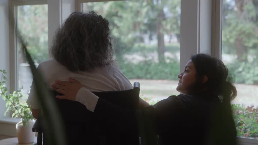 A young dark haired woman wearing a black jacket kneels beside a woman with gray curly hair sitting in a wheelchair affectionately patting her shoulder while looking outside a bright window at a lush green outdoor scene