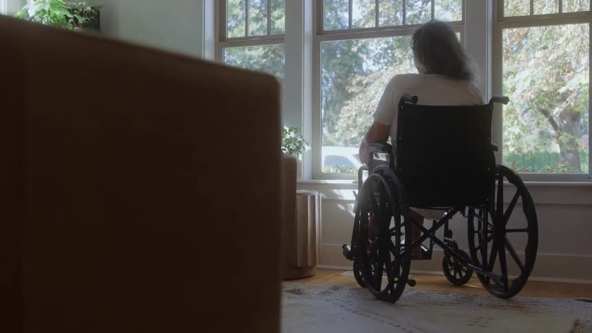 A man with long hair sits in a black wheelchair inside a sunlit room looking out of a large window onto the green trees and neighborhood outside