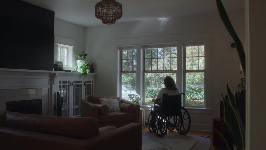 A wide rear view shows a woman sitting alone in a black wheelchair looking out of a large bright window in a cozy living room with leather furniture