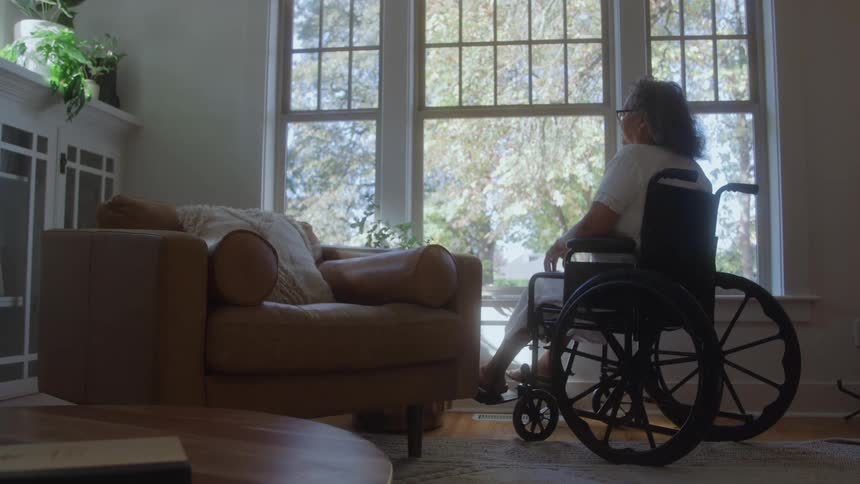 Older woman sitting in a wheelchair looks out the living room window