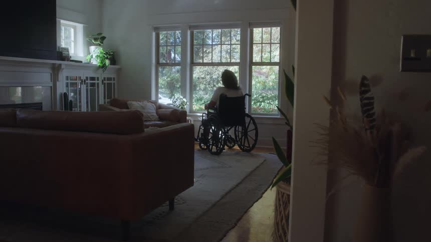 Woman in a wheelchair looking out a sunny window in a living room
