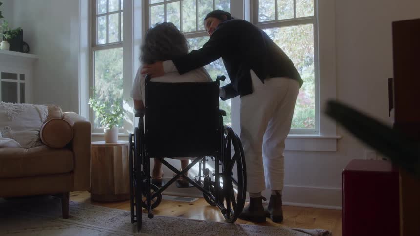 A rear view shows a person in white pants and a dark jacket kneeling to comfort a woman with curly gray hair sitting in a black wheelchair facing a large window in a bright living room