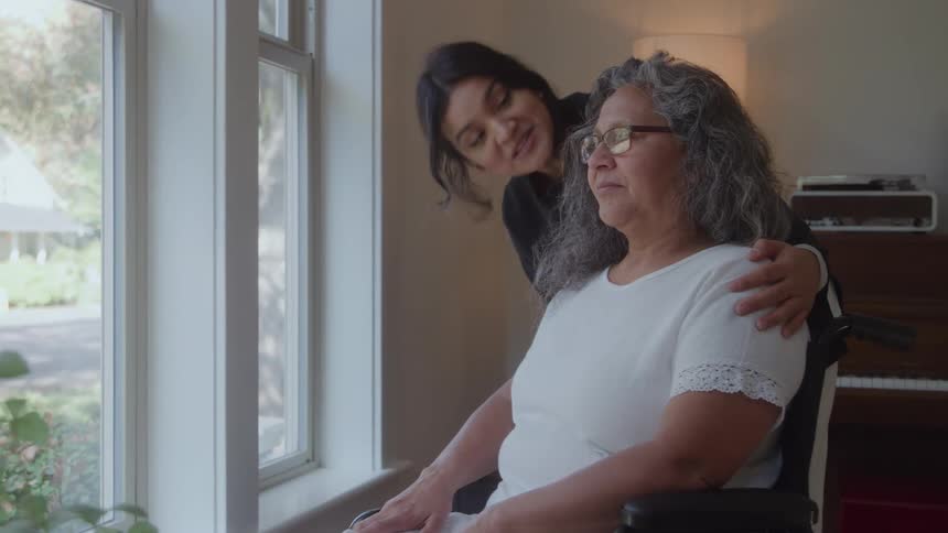 Daughter embraces her mother in a wheelchair by a window indoors
