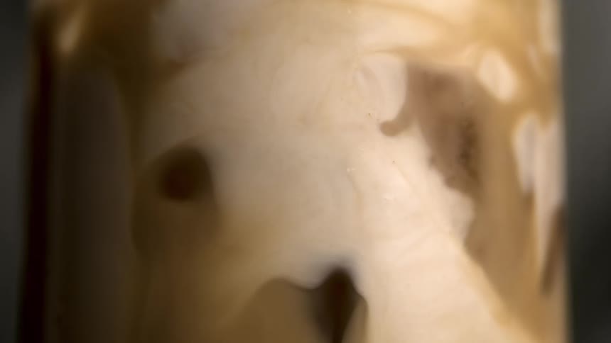 A close abstract view shows light brown and creamy white liquid swirling and blending together inside a clear glass container