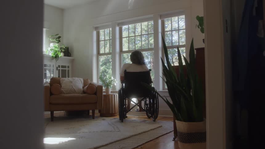 Older woman sits in a wheelchair in her living room and looks out the window