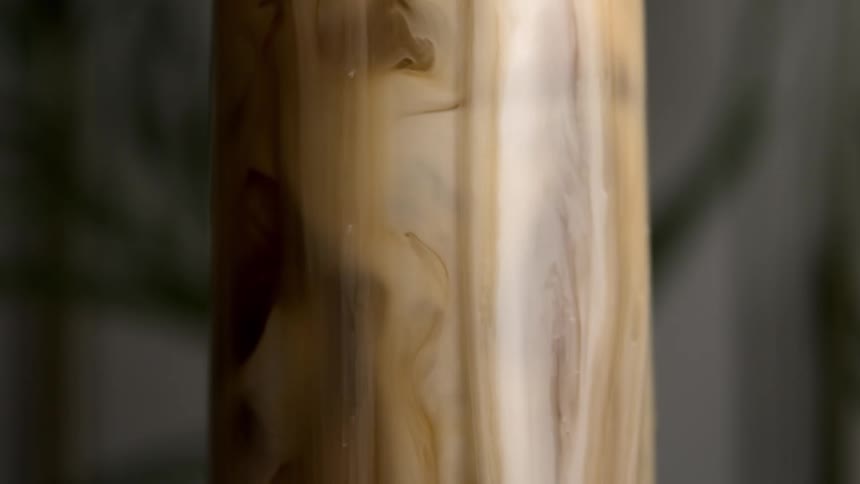 The video is an extreme closeup shot of a milky brown beverage swirling inside a clear glass or cylindrical containerthe two liquids are mixing with a mesmerizing creamy pattern suggesting coffee tea or another layered drink being stirred