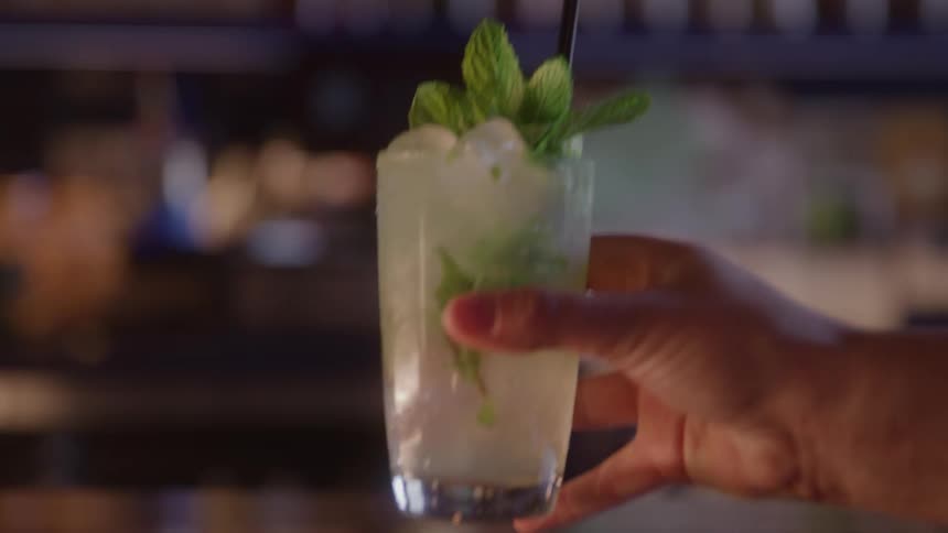 A mans hand reaches for a tall glass containing a refreshing cocktail garnished with ice a straw and a large sprig of fresh mint in a dim bar setting