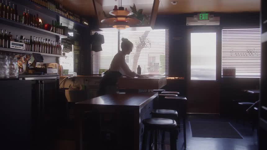 A bartender cleans a bar counter in a dim stylish space lit by sunlight through blinds