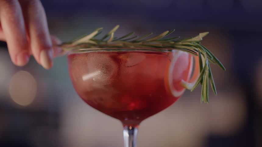 A hand sets a sprig of rosemary on a red cocktail in a coupe glass with a citrus wheel on the rim