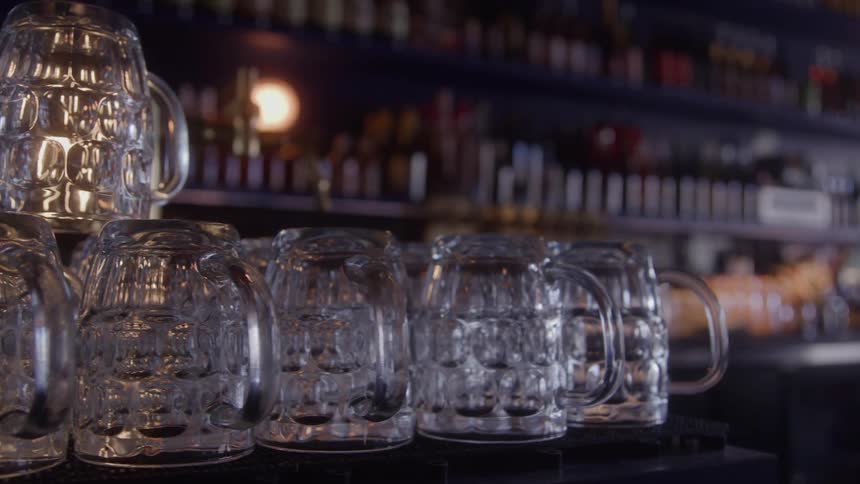 A shot focuses on a row of clean empty textured glass beer mugs lined up on a dark bar surface the background is a dimly lit bar shelf with various liquor bottles and soft outoffocus lights visible