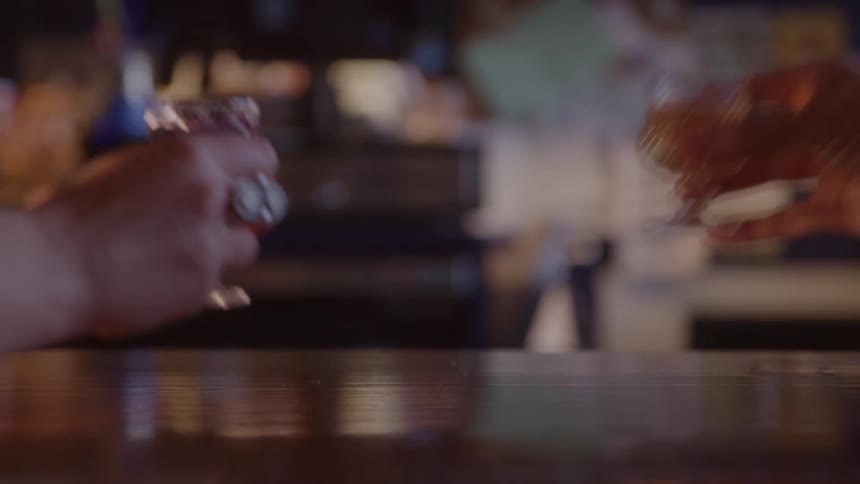 A trio of hands reaches for tiny stemmed glasses of liqueur on a polished bar