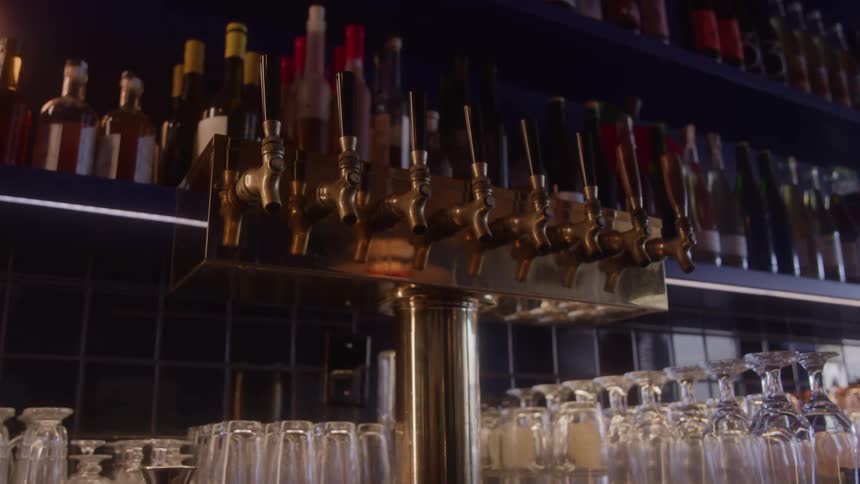 A view of the bar shows a row of golden brass beer taps a collection of clean empty glasses and shelves filled with various bottles in a dimly lit pub