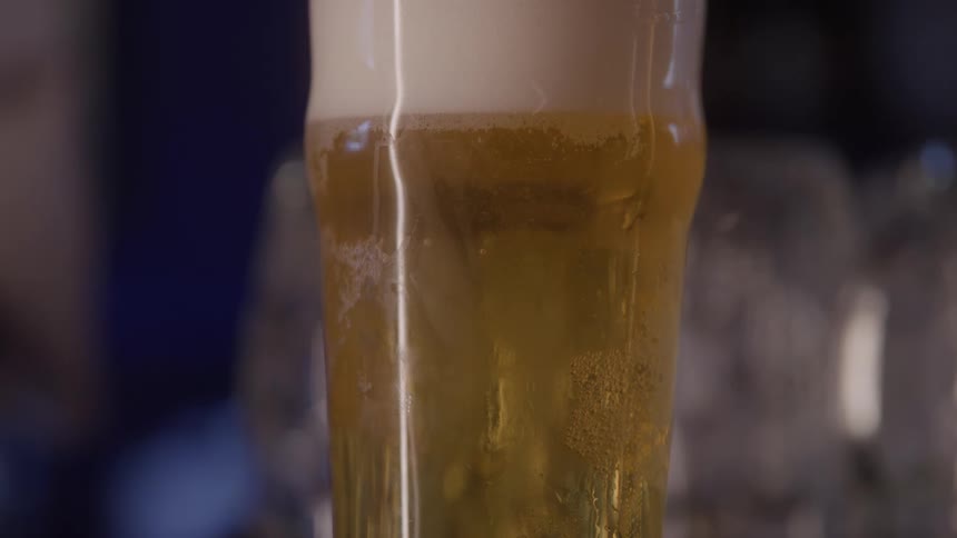 A tall pint glass filled with golden beer sits still showing a layer of white foam and tiny bubbles rising slowly through the liquid