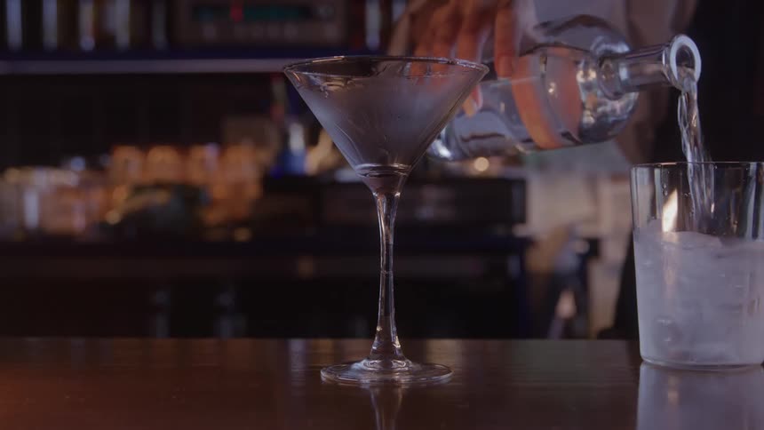A bartender pours clear liquor into an ice filled rocks glass beside an empty chilled martini glass