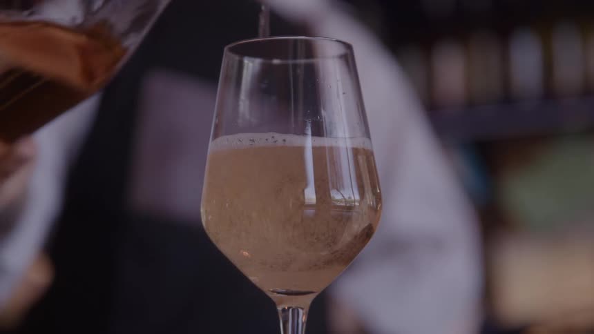 A close view shows sparkling rose colored wine being poured from a glass bottle into a tall clear champagne flute by a person wearing a formal black and white uniform