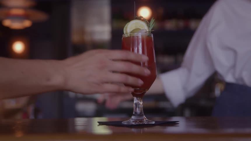 Hand reaching for a red cocktail with lemon and rosemary at a dark bar