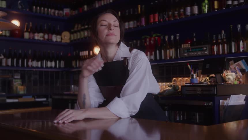 A bartender a woman in a white shirt and dark apron is leaning casually on a long polished wooden bar in a dimly lit cozy establishment the background features shelves lined with numerous dark bottles of wine or liquor creating a warm moody