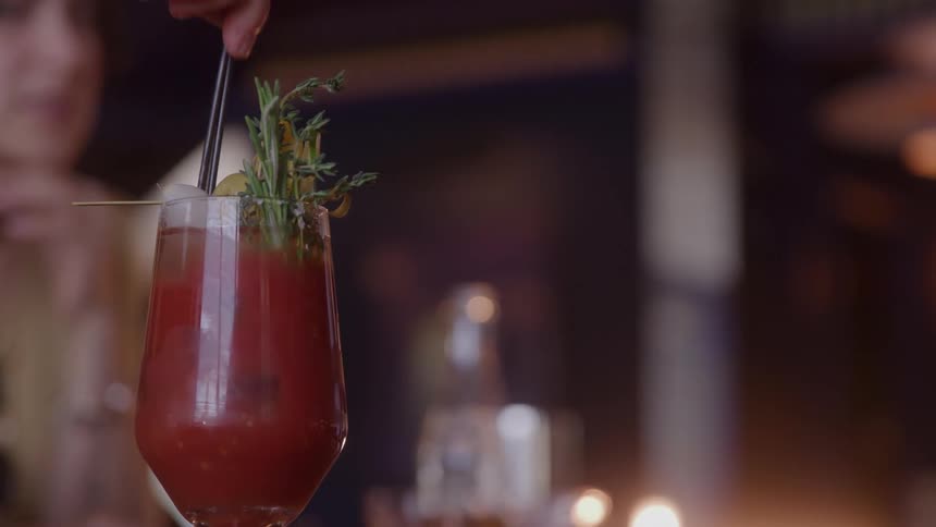 A persons hand using a straw to stir a red drink with some decorative plants on top