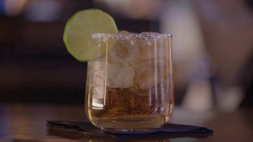 The video shows a closeup of a lowball glass filled with a light brown beverage ice and garnished with a lime wedge on the rimthe rim of the glass is coated with a salt or sugar rim suggesting it is a cocktail like a margarita or a similar