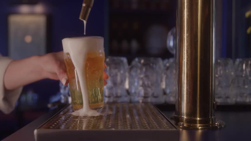 A hand holds a pint glass under a tap as golden beer pours and foams over the rim on a bar counter