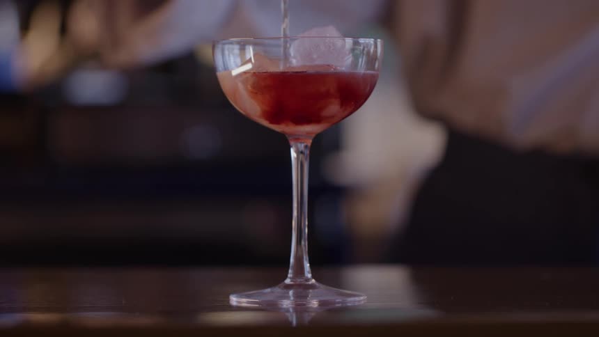 Bartender pouring red cocktail into a coupe glass on a bar countertop