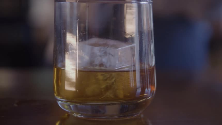 A piece of ice drops into a tumbler glass containing whiskey and the glass is sitting on a reflective dark table surface the background is out of focus
