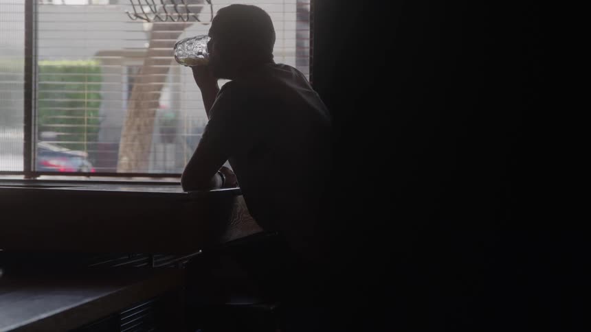A man sitting in a dark bar drinks the rest of his beer then gets up