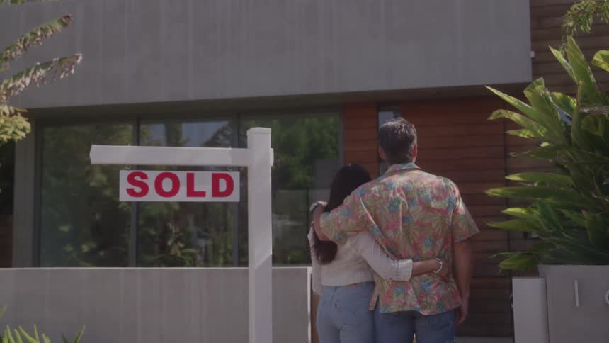 A couple stands arm in arm outside a modern house beside a sold sign