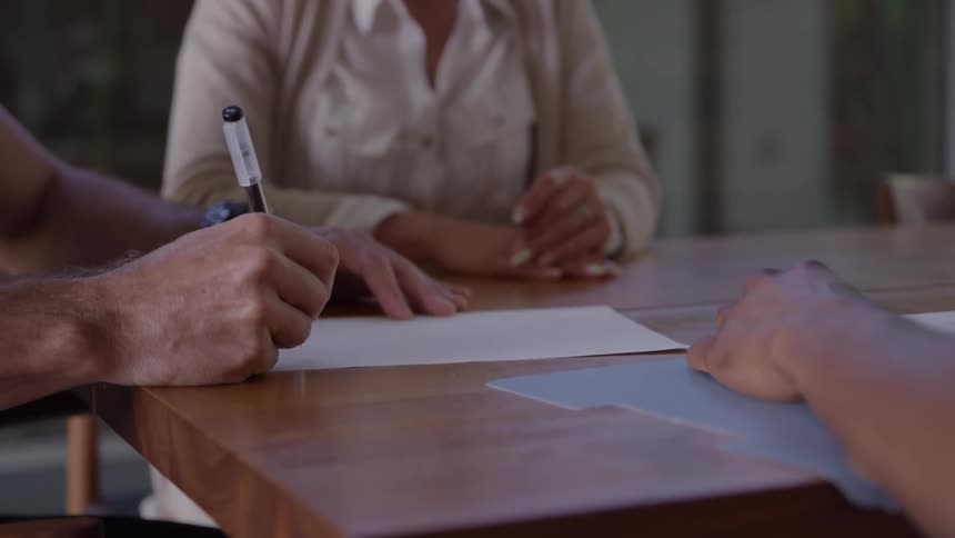 A close view shows a mans arm and hand signing a document with a black pen on a wooden table while a woman in a beige cardigan watches in the background