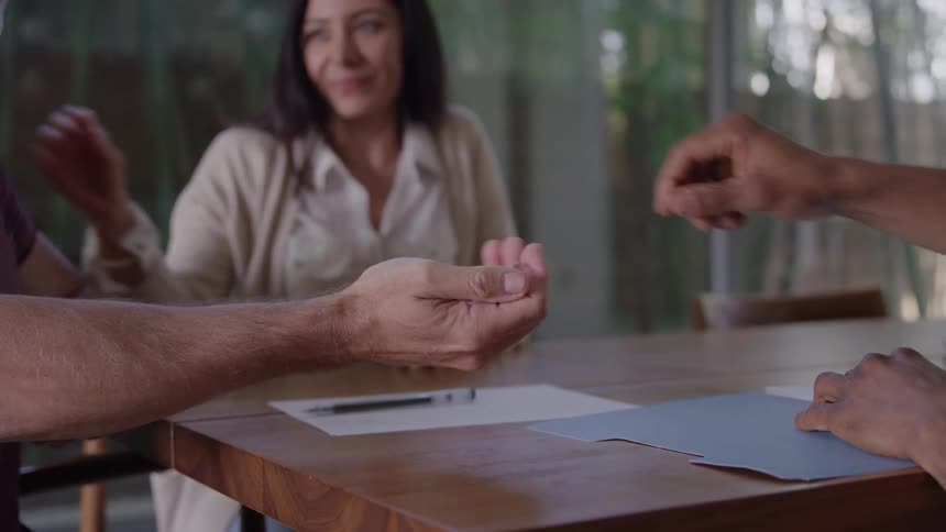 The video shows a group of three people sitting at a wooden table engaged in a business or planning discussiontheir hands are visible pointing to papers and reaching across the table suggesting collaboration and agreement