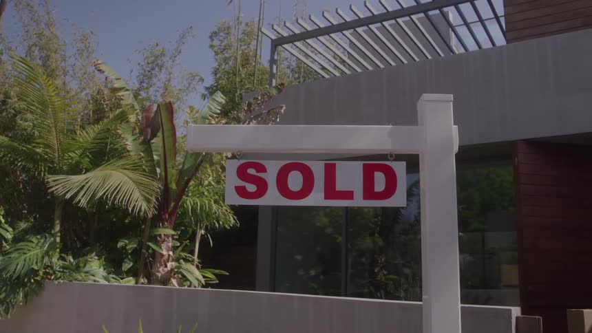A white real estate sign with the word sold in red letters stands in front of a modern home surrounded by lush landscaping under a clear blue sky