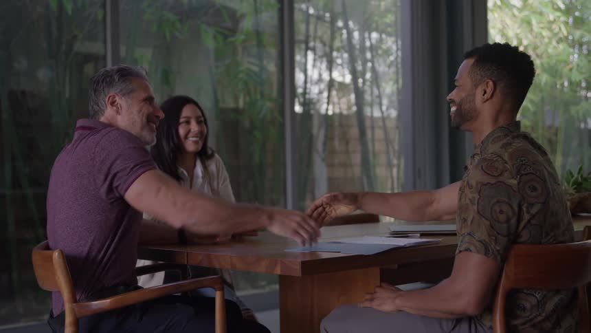 A happy woman in a white sweater and two men in button up shirts sit at a wooden table in a sunlit modern room with large glass windows and shake hands across the table after a successful business meeting