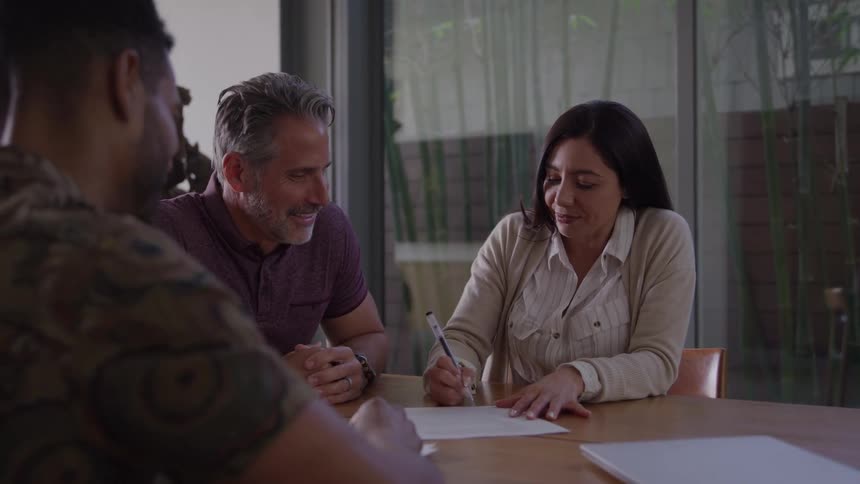 A woman receives a document from a man in the foreground sits next to a man smiles signs the paper with a pen then leans in to hug the man sitting beside her