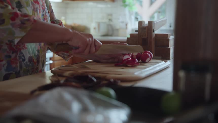 A person slices radishes on a wooden cutting board in a bright kitchen