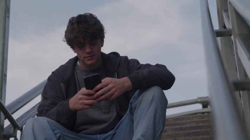 A young man with curly brown hair is sitting alone on a set of concrete stairs outdoors framed by a metal railing on his left he is dressed casually in a dark hoodie and jeans looking down intently as he uses his smartphone