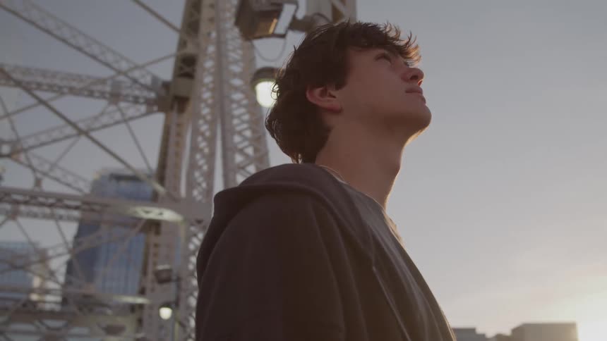 A teenage boy stands outdoors his head tilted upward toward the sky around him theres a large metal structure of a bridge overhead city buildings visible in the distance and the sun setting in the background