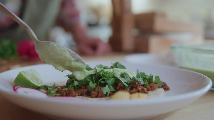 A close view of a taco shows a hand spooning a creamy green sauce over a corn tortilla filled with ground meat and fresh chopped cilantro and garnished with a lime wedge and radish slices
