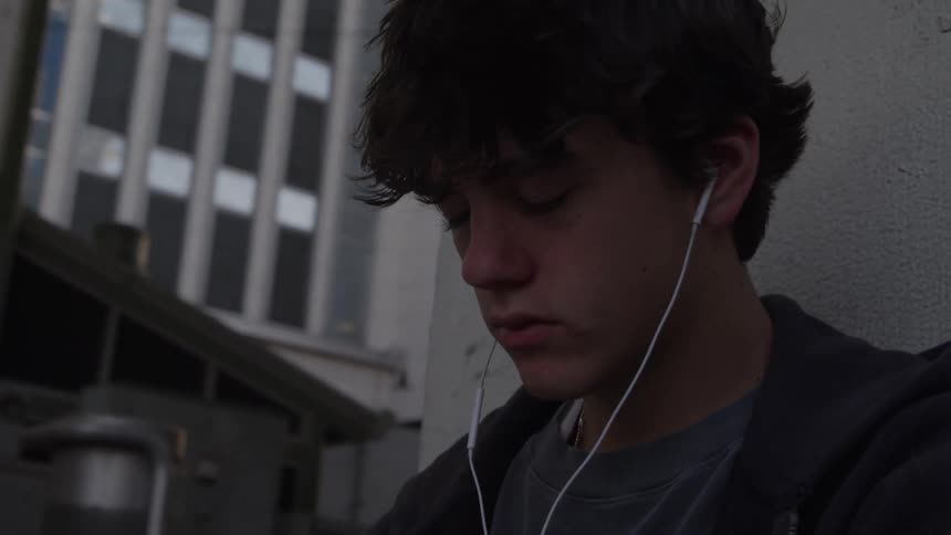 The video is an establishing shot of an urban street in a city focused on a young man sitting alone on the pavementthe man who appears to be listening to headphones is slumped against a concrete wall with a melancholic or weary posture